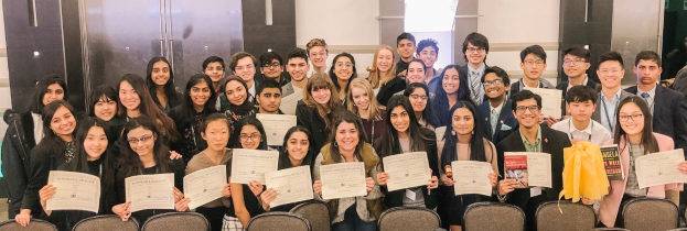 Eine Gruppe von Menschen hält Urkunden in der Hand und posiert für ein Foto in einem Raum mit Stühlen und einer Wand im Hintergrund, wahrscheinlich bei einer Abschlussfeier für das akademische Jahr 2019-2020.