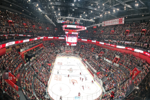 Eine große Indoor-Eishockeyarena mit Zuschauern auf den Rängen, heller Deckenbeleuchtung und einem zentralen Anzeigebildschirm während eines Spiels.