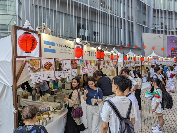Eine Gruppe von Menschen auf einem Food-Festival in Japan versammelt sich um einen Tisch mit Essen, einige halten Gläser und tragen Taschen, während andere die Ausstellungsstücke inspizieren.