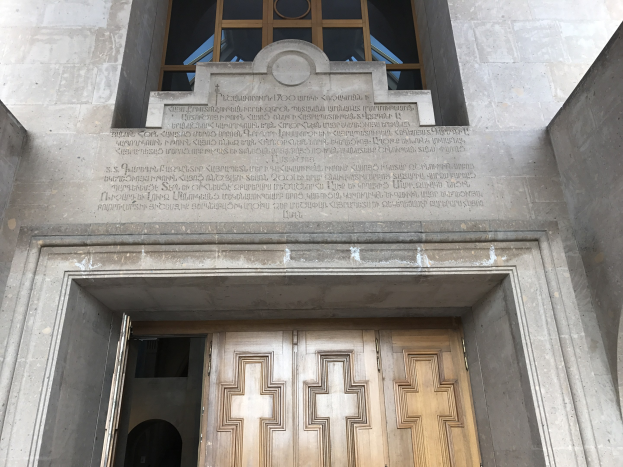 Große hölzerne Tür vor einem steinernen Gebäude mit einem Glasfenster oben und einer Inschrift an der Wand, wahrscheinlich ein Holocaust-Mahnmal.