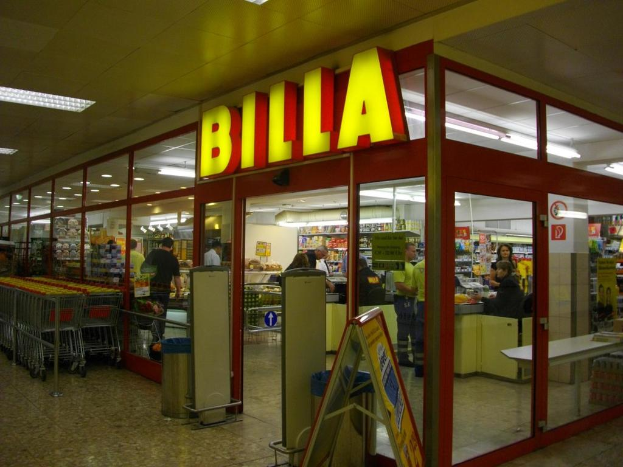 Ein Supermarkt mit einem Eingang in der Mitte, Mülltonnen daneben, einer Reihe von Einkaufswagen links, Deckenbeleuchtung und einigen Menschen, die Produkte anschauen.