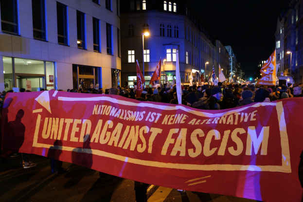 Eine Gruppe von Menschen marschiert mit einem Banner, auf dem "Einheit gegen Faschismus" steht, eine Straße entlang, mit Gebäuden, Laternenmasten und Schildern an der Straße und einem sichtbaren Himmel im Hintergrund.