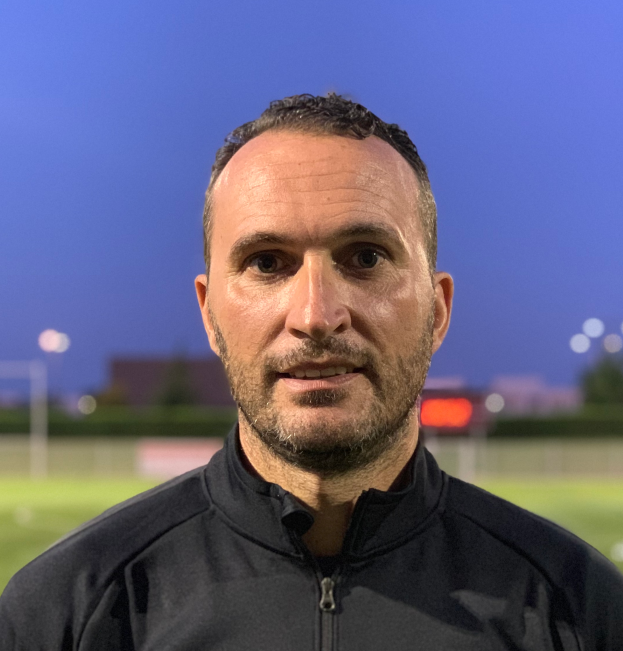 Ein Mann in einer schwarzen Jacke mit Logo steht auf einem Fußballfeld und sieht aus wie ein Trainer mit einem unscharfen Hintergrund.