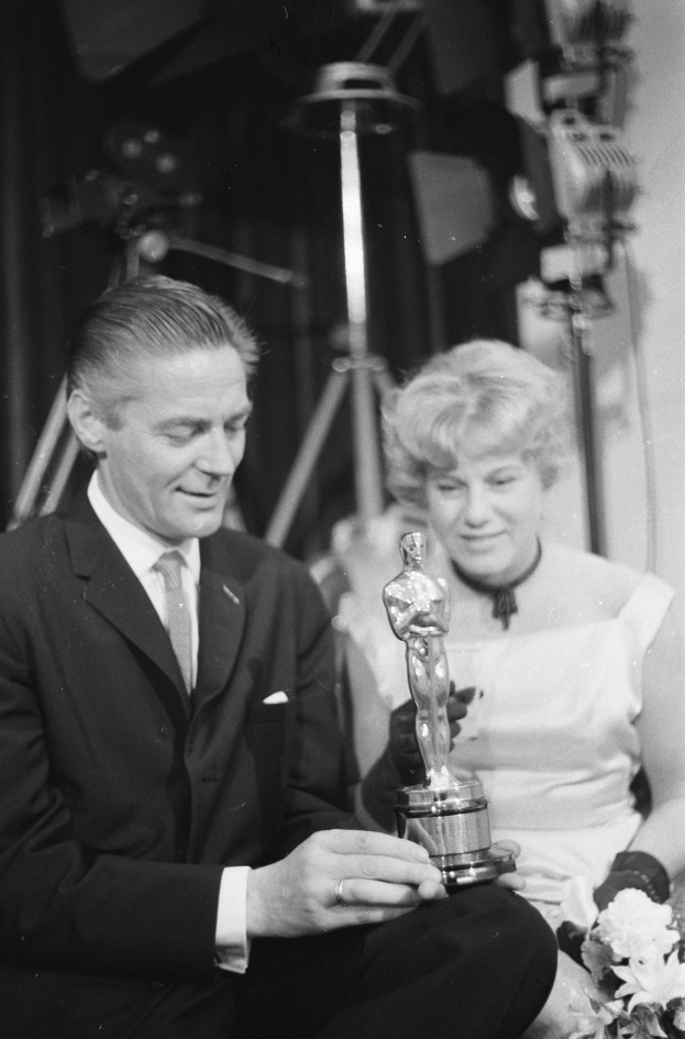 Ein Schwarz-Wei├č-Bild eines formell gekleideten Mannes, der eine Oscar-Statue hölt und eine Frau, die einen Blumenstrau├č hölt, nebeneinander sitzen.