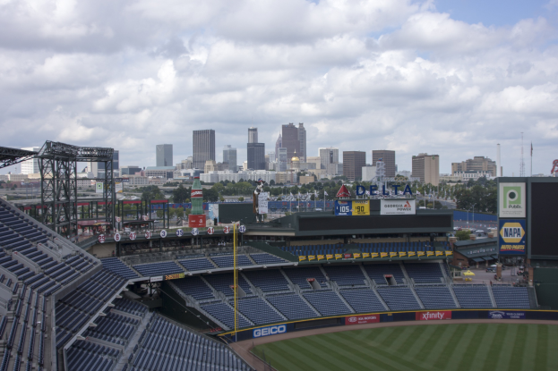 Baseballstadion mit einer Stadtkulisse im Hintergrund, gefüllt mit Stühlen, Pfosten und Brettern, auf Gras mit Bäumen und Gebäuden in der Ferne unter einem bewölkten Himmel.