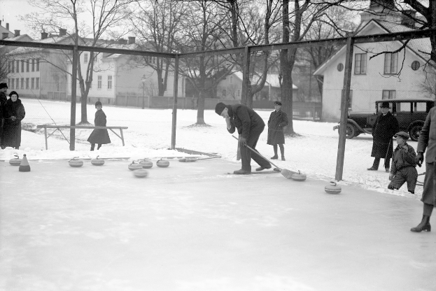 Schwarzes Bild von Menschen, die Curling auf einem Outdoor-Eisstadion spielen, umgeben von einem Zaun, einer Bank, Bäumen, Gebäuden und einem geparkten Fahrzeug im Hintergrund.