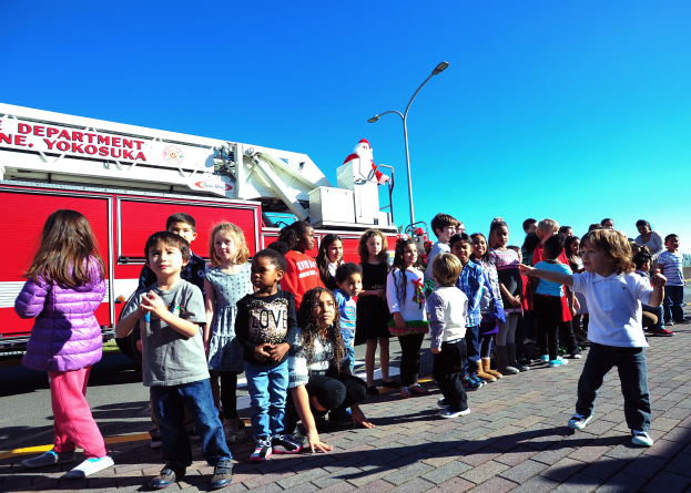 Kinder vor einem Feuerwehrauto mit der Aufschrift 'Yokosuka Fire Department's Christmas Parade' auf einer Straße mit Laternenmästen, Bäumen und einem klaren blauen Himmel.
