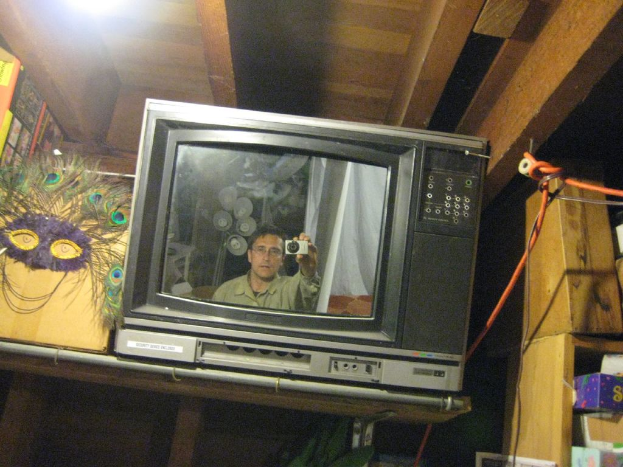 Ein Fernseher mit einer Person umgeben von Pfauenfedern, B├╝chern und Kisten.