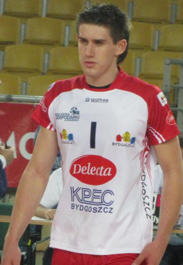 Ein Mann in einem rot-weißem Handballtrikot steht auf einem Feld, umgeben von Zuschauern, mit einem roten Banner im Hintergrund, auf dem "Krasnoyarsk vs Bydgoszcz" steht, und sieht entschlossen aus.
