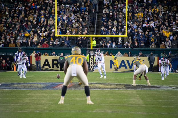 Fußballspieler in Sportbekleidung und Helmen bei einem Spiel auf einem Feld mit Zuschauern im Hintergrund, Stadionstrukturen und sichtbaren Schildern.