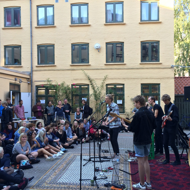 Eine Gruppe von Menschen sitzt auf dem Boden und spielt live mit Musikinstrumenten, Mikrofonen und verstreuten Gegenständen vor einem Gebäude in Berlin.