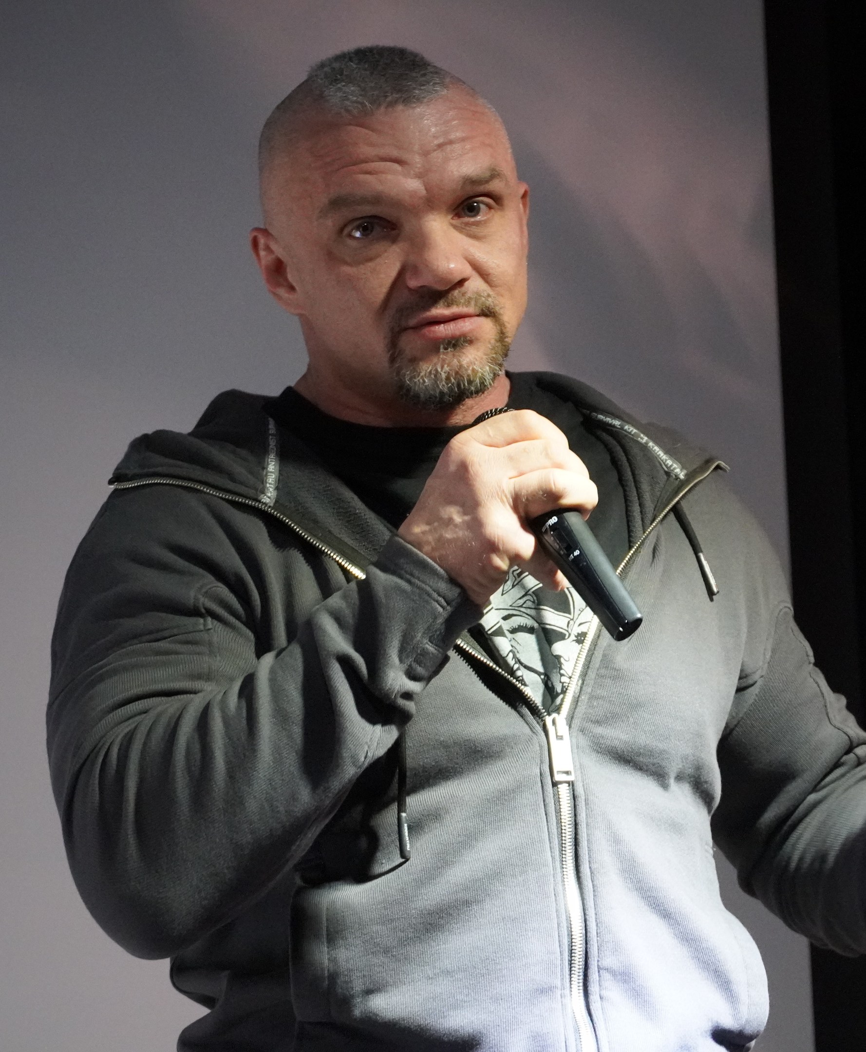 Ein Mann in einer schwarzen Kapuzenjacke und einem schwarzen Anzug mit weißem Hemd hält ein Mikrofon und steht selbstbewusst vor einer Wand, wobei er wie ein Komiker wirkt.