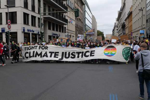 Eine Gruppe von Menschen marschiert mit einem Banner, auf dem "Kampf für Klimagerechtigkeit" steht, eine Straße entlang, mit Gebäuden, Schildern, Verkehrszeichen und Bäumen an der Straße unter einem bewölkten Himmel.