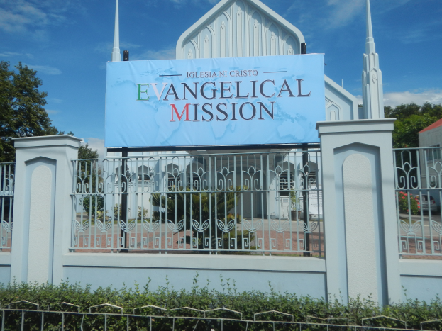 Eine Kirche mit einem Schild "Evangelische Mission" ist von einem Metallzaun umgeben, Pflanzen, Tälern und einem Haus mit einem Dach unter einem bewölktem Himmel.