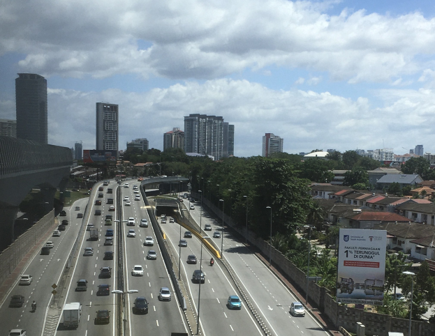 Eine belebte Autobahn mit hohen Gebäuden, eine Brücke auf der linken Seite, Laternenpfähle, Bäume und ein bewölkter Himmel.
