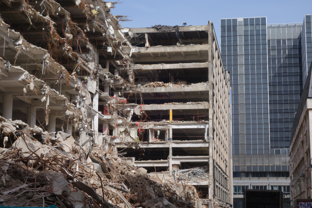 Ein großes abgerissenes Gebäude inmitten einer Stadt, umgeben von anderen Gebäuden, Straßenlaternen, Namensschildern, Kraftfahrzeugen, Bäumen und einem klaren blauen Himmel.