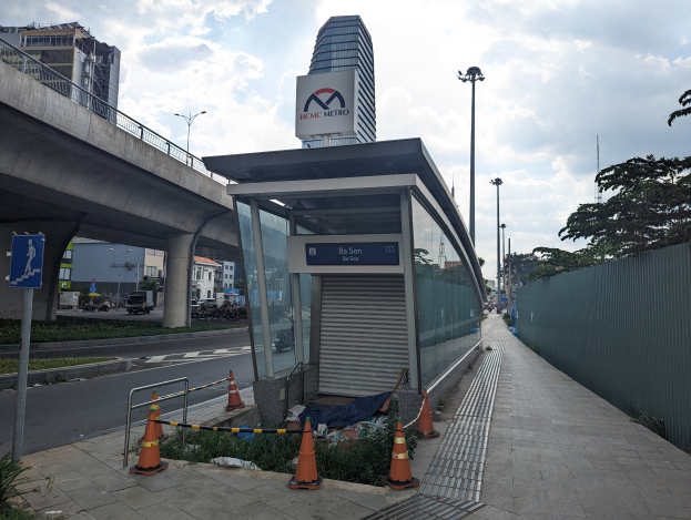 Eine Bushaltestelle am Straßenrand neben einem hohen Gebäude, umgeben von Absperrbaken, Pflanzen, Gras, einem Zaun, Fahrzeugen, einer Tafel, Straßenlaternen, einer Brücke, Gebäuden mit Fenstern und einem bewölkten Himmel.