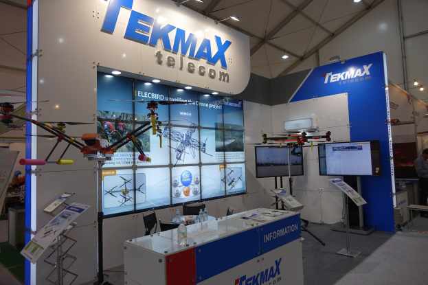 Ein Messestand für Tekmax Telecom mit einer großen Wandanzeige, einem Tisch mit Produkten, zwei Fernsehern, Infotafeln, Deckenleuchten und einigen Menschen.