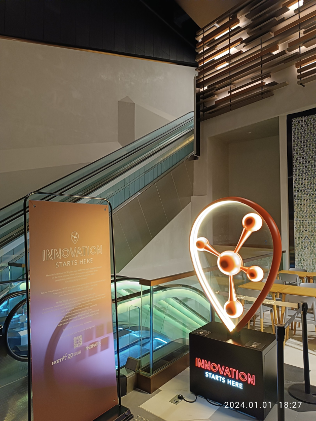 Großes Schild in einem Gebäude mit einer Rolltreppe im Hintergrund, Tische und Stühle auf der rechten Seite und eine Tafel mit der Aufschrift "Innovation beginnt hier" auf der linken Seite, die auf die HSTP20 Expo hinweist.