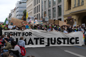 Eine große Gruppe von Menschen marschiert mit einem Banner 'Kämpfen für Hassjustiz' die Straße entlang, einige tragen Masken und halten Schilder oder Taschen, mit Gebäuden, Bäumen und einem klaren blauen Himmel im Hintergrund.