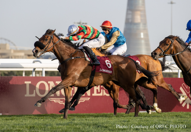 Drei Jockeys auf Pferden, die auf einer grünen Rennbahn mit einer Texttafel und einem Turm im Hintergrund unter einem klaren blauen Himmel galoppieren.