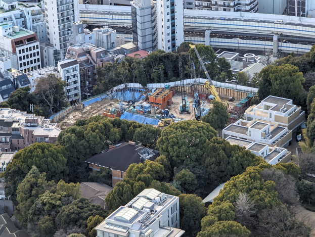 Luftaufnahme einer städtischen Baustelle mit Gebäuden, Bäumen, Fahrzeugen, Kränen und einer Brücke im Hintergrund, die verstreute Werkzeuge und Materialien zeigt.