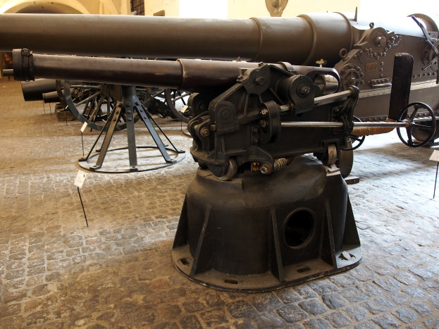 Eine große Gatling-Kanone mit langem Lauf und seitlichem Griff ist in einem Museum ausgestellt, umgeben von Bodenständen, mit einer fensterumgebenen Wand im Hintergrund, die an eine Burg erinnert.