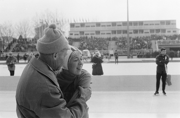 Ein Mann und eine Frau umarmen sich auf einem Eisstadion vor einem Publikum, wobei der Mann eine Jacke und eine Mütze trägt und die Frau lächelt, während andere Eisläufer, Bäume, Gebäude und ein Pfahl im Hintergrund zu sehen sind.
