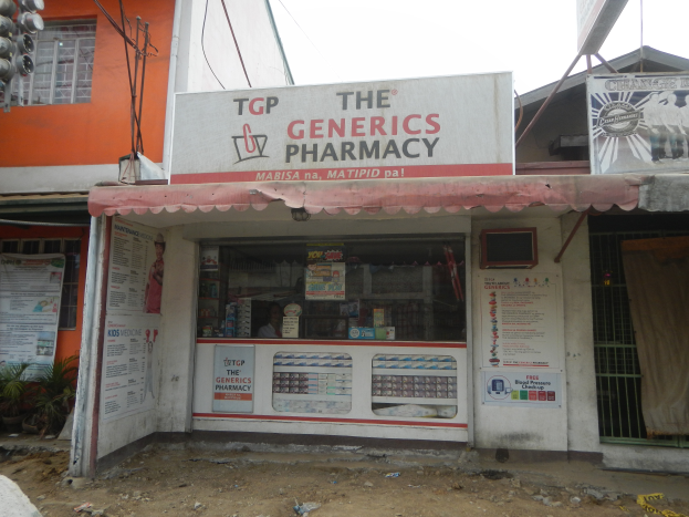 Eine Apotheke namens "Die Generika Apotheke" umgeben von Gebäuden, Strommasten, Werbetafeln, Zimmerpflanzen, Kraftfahrzeugen auf der Straße, unter einem klaren blauen Himmel.