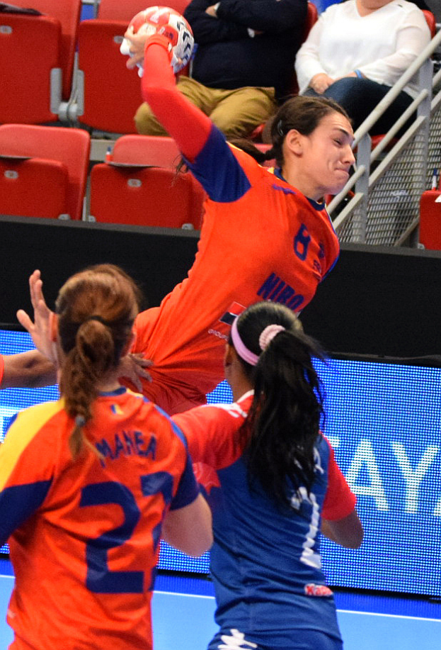Eine Gruppe von Frauen, die in einem Stadion Handball spielen, wobei eine den Ball hält und Zuschauer im Hintergrund sitzen.