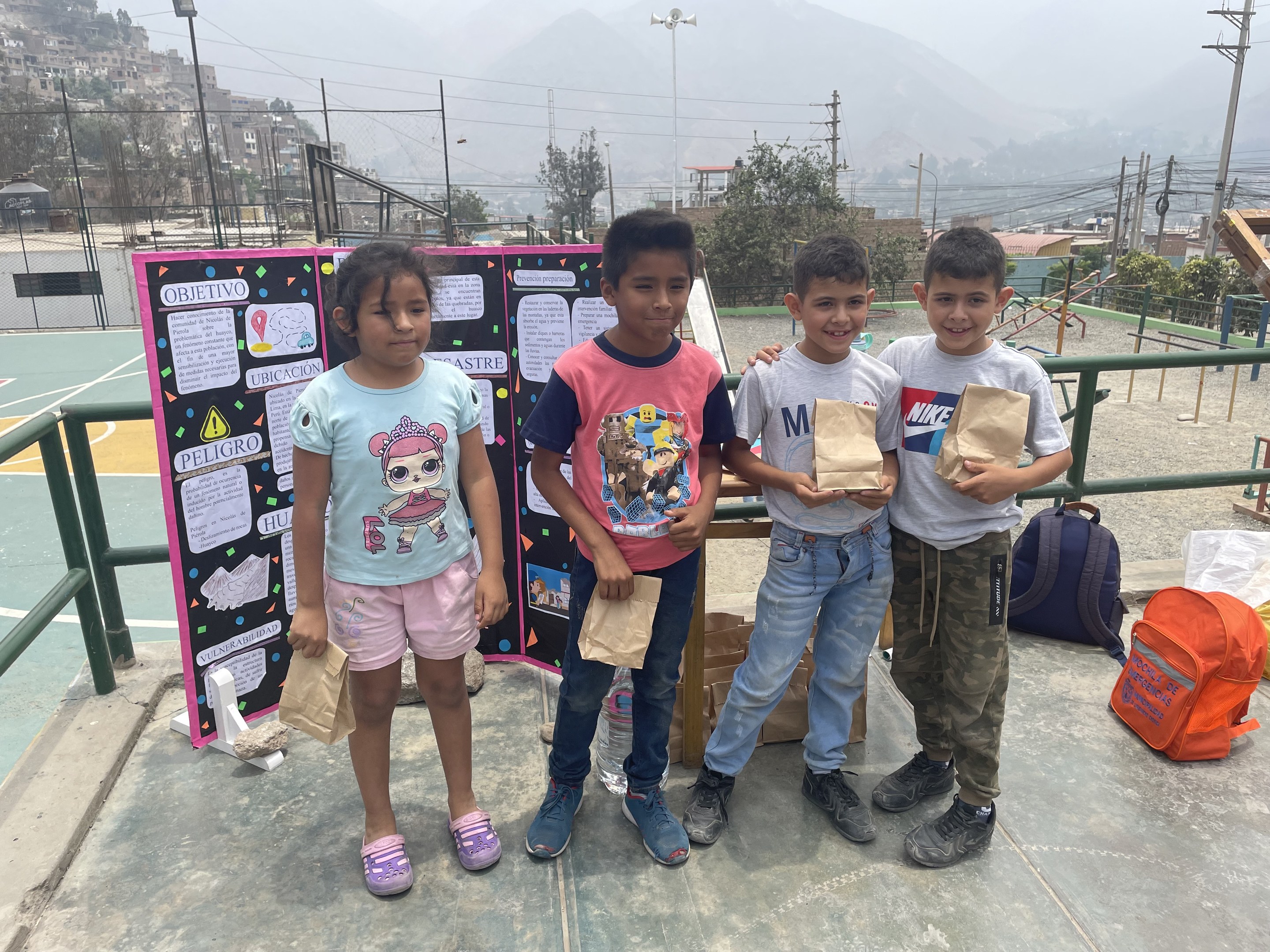 Gruppe von Kindern auf einem Basketballfeld mit Papiertüten stehend, Tüten und Gegenstände rechts und eine Tafel mit Text links, vor einem Hintergrund aus Zaun, Pfosten, Laternen, Bäumen, Gebäuden, Hügeln und Himmel.