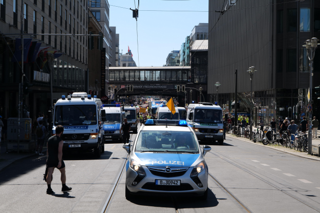 Eine Gruppe von Polizeiwagen fährt eine Straße mit hohen Gebäuden, Fußgängern auf dem Gehweg, parkenden Fahrrädern und Laternenmasten entlang, mit einer Brücke und einem klaren blauen Himmel im Hintergrund.