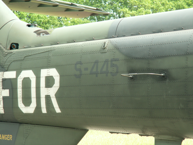 Nahaufnahme eines Militärhubschraubers mit dem Wort "FOR" auf der Seite, vor einem Baumhintergrund.