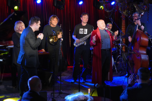 Eine Gruppe von Menschen auf einer Bühne mit Musikinstrumenten, Mikrofonen und einer Kerze, die vor einem sitzenden Publikum spielt.