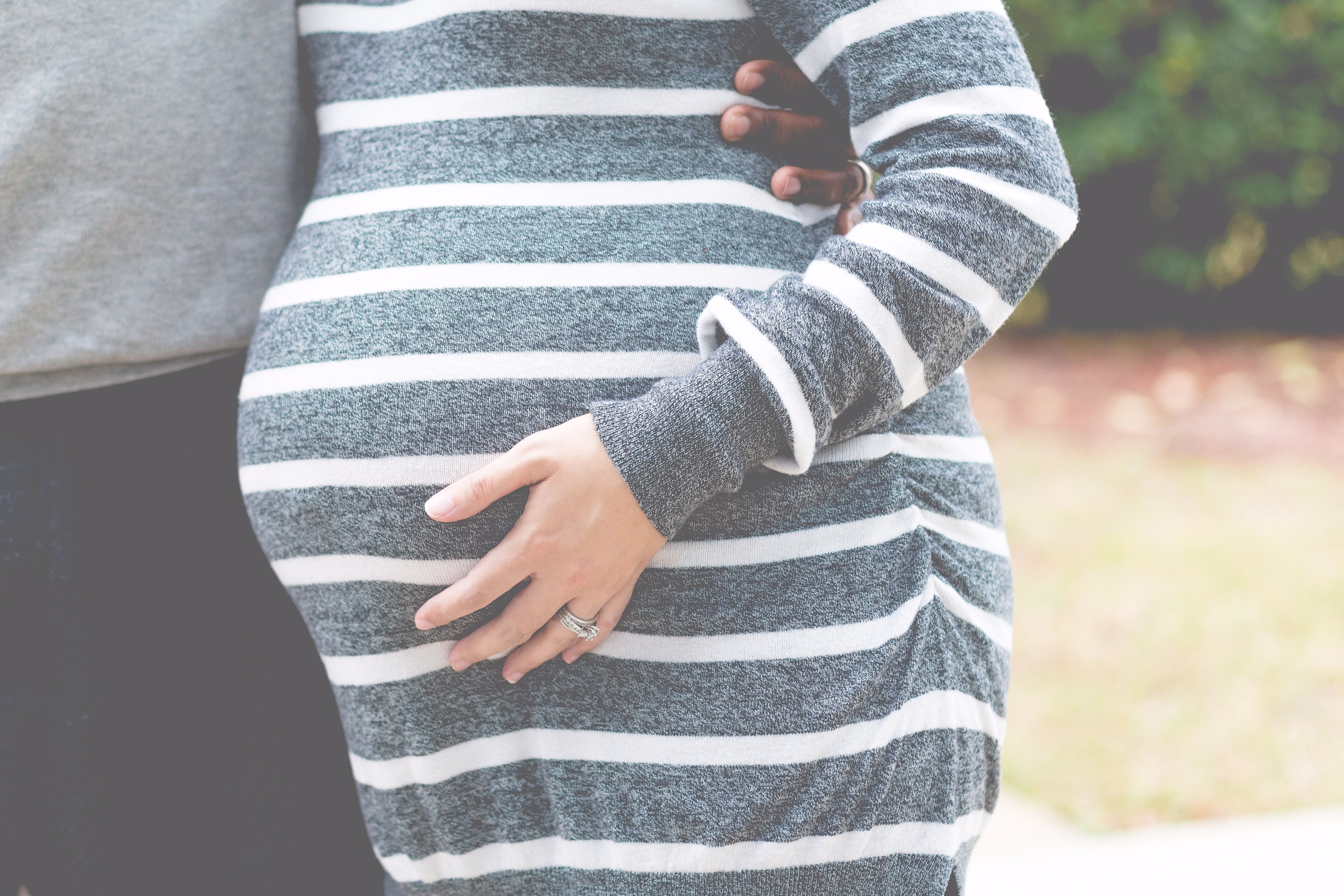 Schwangeres Paar in Umstandskleidung, das zusammen steht und den Bauch hält, umgeben von Bäumen.