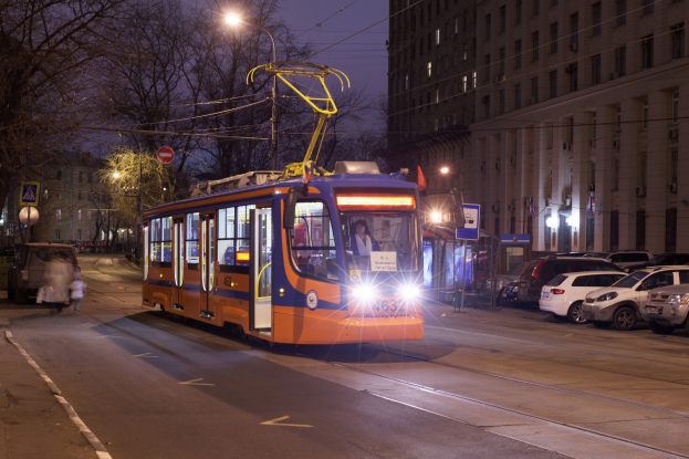 Orange und blaues Tram auf einer Stadtstraße bei Nacht mit Passagieren im Inneren, parkende Autos auf der rechten Seite, Gebäude und Bäume im Hintergrund und beleuchtete Straßenlaternen und Lichtmasten unter einem klaren Himmel.