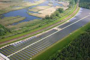 Eine Luftaufnahme eines Solarparks mit zahlreichen Solarmodulen auf einem Feld, umgeben von Bäumen, Gras und Wasser, mit einem Zug, der auf einem Bahnsteig in der Nähe fährt.