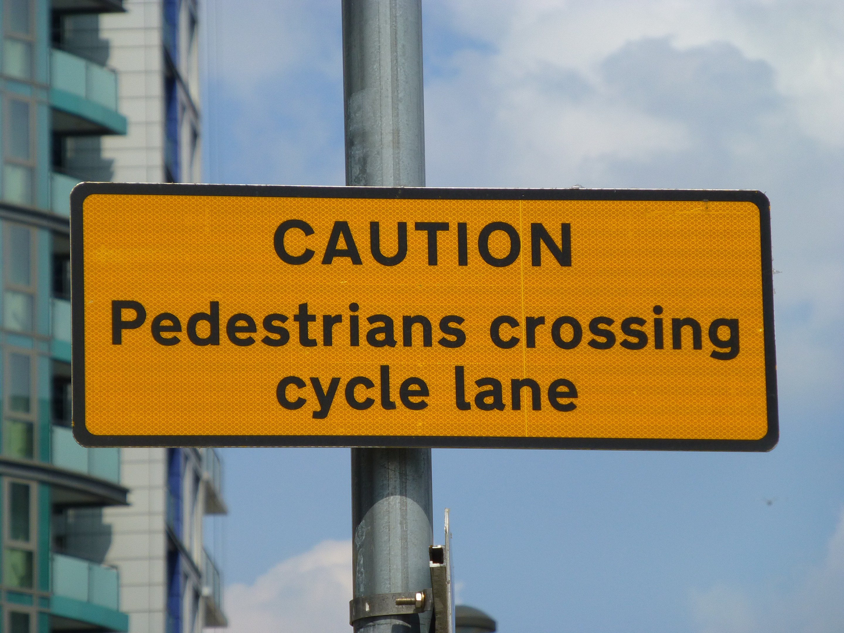Gelbes Warnschild an einem Pfosten mit der Aufschrift "Fußgänger überqueren Radweg" vor einem Gebäude unter einem klaren blauen Himmel.