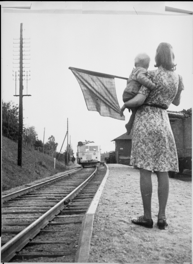 Eine Frau in einem Kleid und Schuhen steht auf den Bahnschienen und hält ein Baby, während ein Zug im Hintergrund näherkommt; Pfähle und Bäume sind links zu sehen und der Himmel ist oben zu sehen.