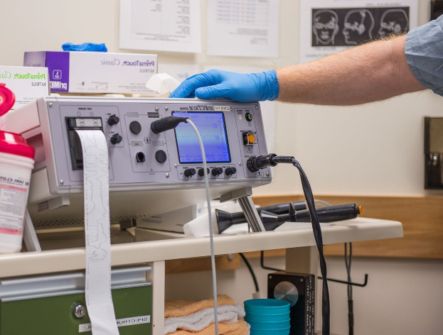 Eine Person in blauen Handschuhen arbeitet an einem medizinischen Gerät auf einem mit verschiedenen Gegenständen übersäten Tisch, mit Papieren an der Wand in einer Krankenhausumgebung.