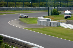 Ein Rennwagen fährt auf einer Rennstrecke mit einem Polizeiwagen im Hintergrund, umgeben von Gras, Pflanzen, Bäumen und Zäunung, mit einem Fahrzeug auf der rechten Seite und Text unten.