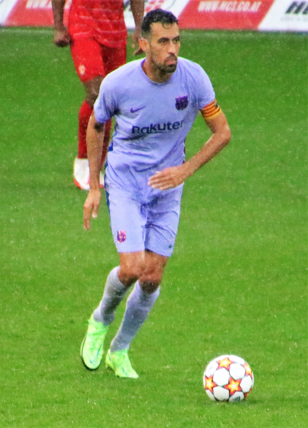 Ein Fussballspieler in Sportbekleidung und -schuhen ist im Einsatz auf dem Feld, wobei eine Tafel mit Text im Hintergrund sichtbar ist.