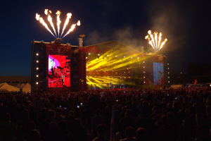 Eine große Menschenmenge versammelte sich auf einem Musikfestival, mit einer Bühne in der Mitte, auf der Musiker im Scheinwerferlicht spielten und von Fahnen umgeben waren, Zelte links daneben und ein dunkler Himmel mit Feuerwerk im Hintergrund.