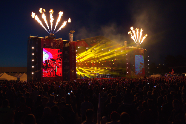 Eine große Menschenmenge versammelte sich auf einem Musikfestival, mit einer Bühne in der Mitte, auf der Musiker im Scheinwerferlicht spielten und von Fahnen umgeben waren, Zelte links daneben und ein dunkler Himmel mit Feuerwerk im Hintergrund.
