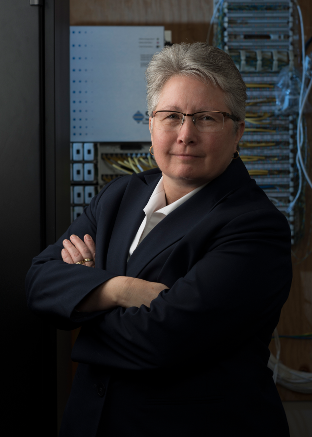 Eine Frau in einem Business-Outfit und Brille lächelt vor einem Server, mit Kabeln und einer Platte an der Wand dahinter.