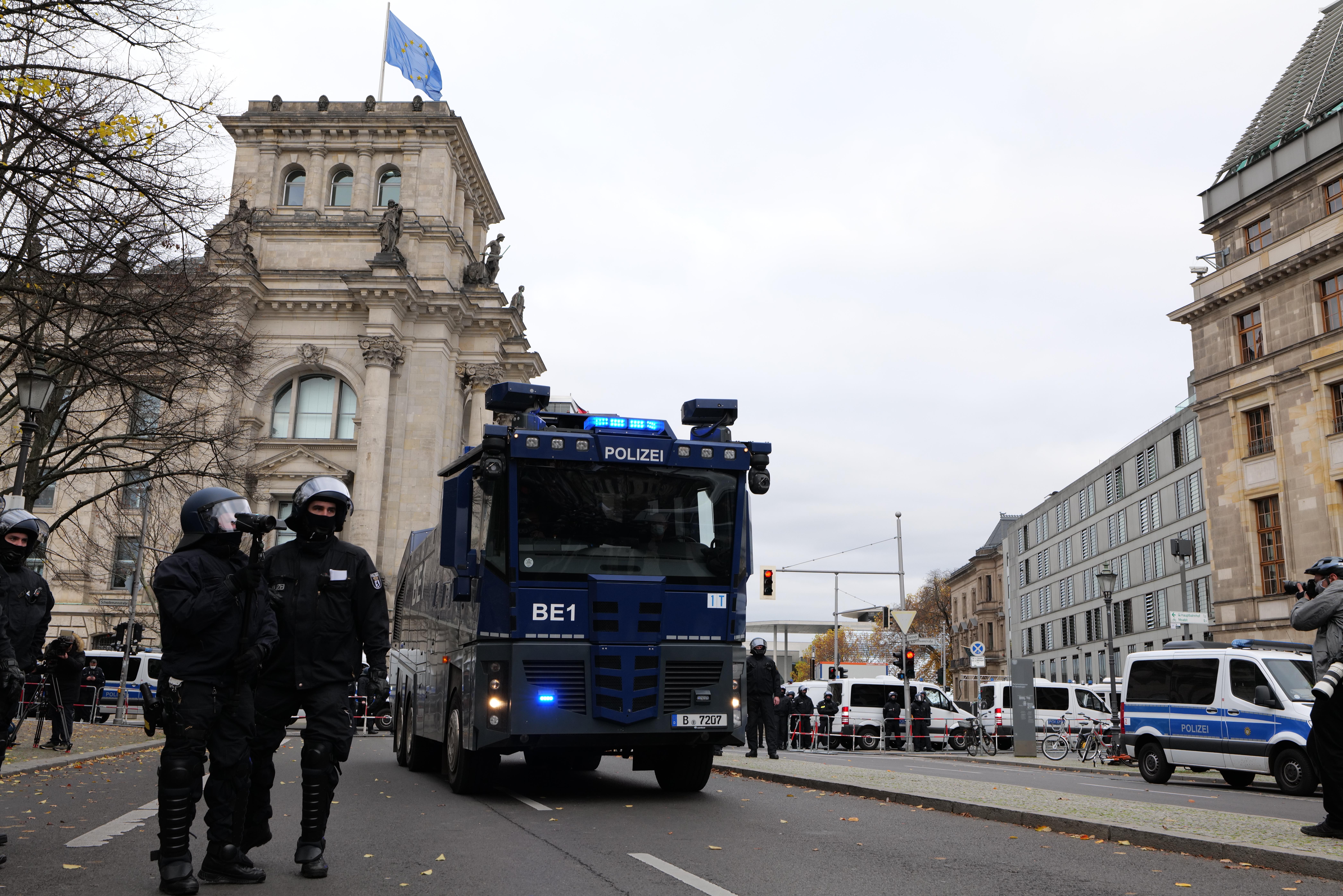 Eine Gruppe von Polizisten in Helmen und mit Waffen in der Hand steht vor einem Gebäude mit Fenstern, Säulen, Bögen und einer Fahne oben, mit Fahrzeugen auf der Straße, Bäumen, Laternenpfählen und einem bewölkten Himmel im Hintergrund.