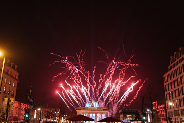 Eine belebte Stadtstraße am Silvesterabend in Berlin, voller Menschen, Fahrzeuge und festlicher Beleuchtung von Gebäuden und Feuerwerk am Himmel.
