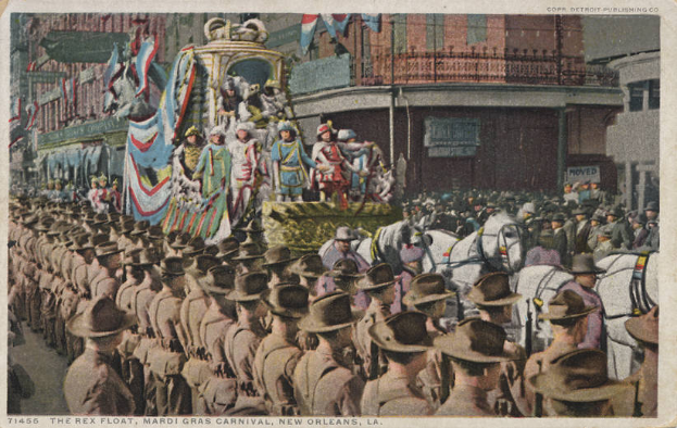 Eine alte Postkarte zeigt einen Mardi Gras-Umzug in New Orleans mit einem Float, der mit Fahnen und Gegenständen geschmückt ist, Menschen zu Pferd und Zuschauern am Straßenrand sowie im Hintergrund Gebäude mit Fenstern und Geländern. Unten steht der Text "The Rex Float, Mardi Gras Carnival, New Orleans".