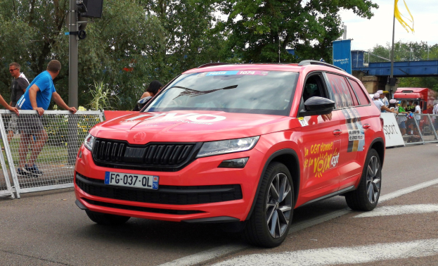Roter Wagen fährt auf einer Straße neben einer Menge, einige tragen Kappen, mit Maschendrahtzaun links, einer Verkehrsampelstange, Bäumen, einer Brücke, einem Fahnenmast und einem bewölkten Himmel im Hintergrund.
