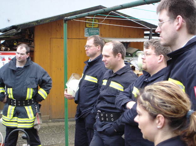 Eine Gruppe von Männern in blauen Feuerwehranzügen, die nach links schauen und vor einem kleinen Holzschuppen und Rohrrahmen stehen.